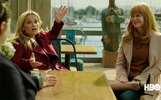 two women sitting at a table talking to each other while another woman holds her hand up in the air