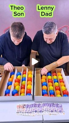 two men sitting at a table with an open box filled with colored balls and money