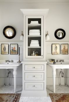 a white bathroom with two sinks and a cabinet in it's center, surrounded by pictures on the wall