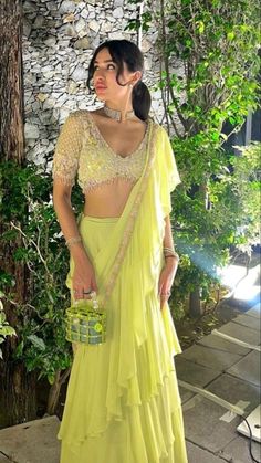 a woman in a yellow sari standing next to a tree and stone wall with greenery behind her