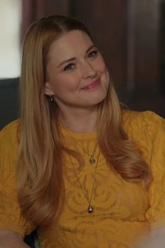 a woman with long blonde hair wearing a yellow top sitting down and smiling at the camera