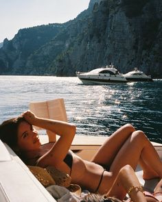 a woman laying on top of a boat next to a large body of water with boats in the background