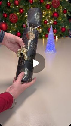 a person holding a black wrapped present in front of a christmas tree with red and gold ornaments