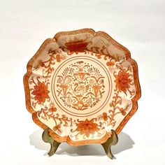 an orange and white plate with flowers on the bottom is sitting on a stand in front of a white background