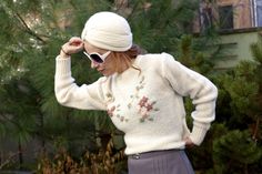 a woman wearing sunglasses and a white knitted sweater stands in front of some pine trees