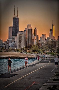 people are walking along the water in front of some tall buildings and skyscrapers at sunset