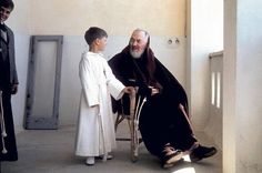 an old man sitting in a chair next to a little boy and another person standing