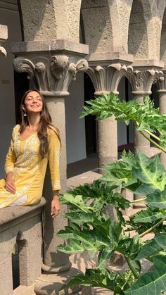 a woman sitting on a ledge next to a green plant and some stone pillars with arches in the background