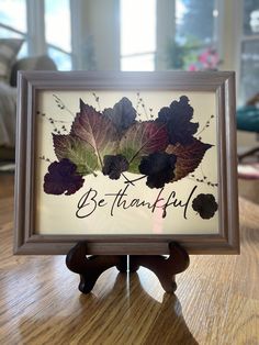 a wooden table topped with a frame filled with leaf pictures and writing that says, be thanful