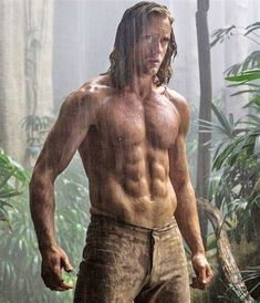 a man with long hair and no shirt standing in the jungle