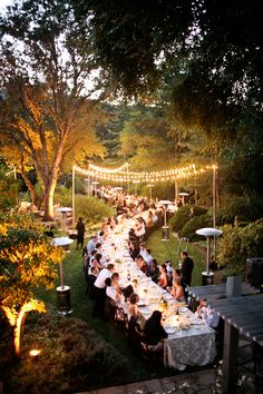 a large group of people sitting at long tables in the middle of a park with lights strung over them