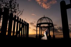 a bride and groom standing in front of a gazebo at sunset