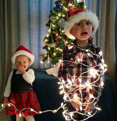 two young children standing next to each other near a christmas tree with lights on it