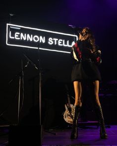 a woman standing in front of a microphone on stage with a neon sign behind her