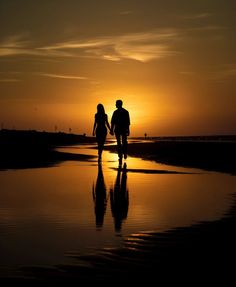 two people walking on the beach at sunset with their reflection in the wet sand as the sun sets