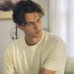 a young man sitting on top of a bed next to a white wall and window