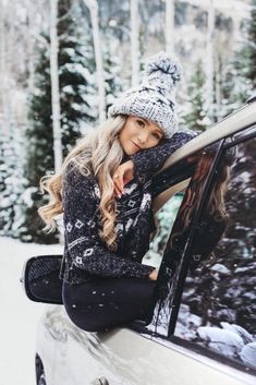 a woman sitting on the hood of a car in the snow wearing a hat and scarf