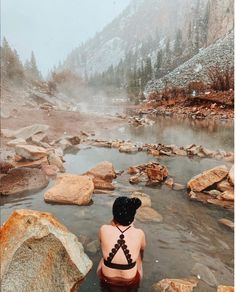 a woman sitting in the middle of a body of water with rocks and trees around her