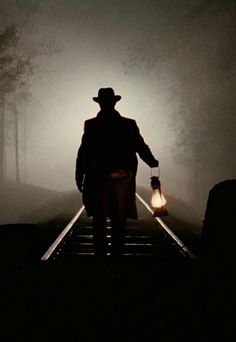 a man in a hat and coat walking down train tracks with a lantern on his hand