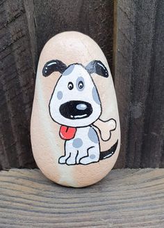 a painted rock with a dog on it sitting next to a wooden fence and holding a red frisbee in its mouth