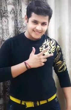 a young man wearing a black shirt and yellow belt giving the thumbs up sign while standing in front of a curtain