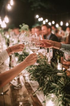 people are sitting at a table with wine glasses in their hands and greenery on the table