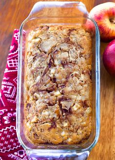 an apple pie in a glass baking dish next to two apples on a wooden table