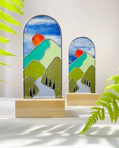 two stained glass windows sitting next to each other on top of a wooden stand in front of a plant