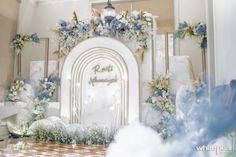 a wedding arch decorated with blue flowers and greenery in front of a white backdrop