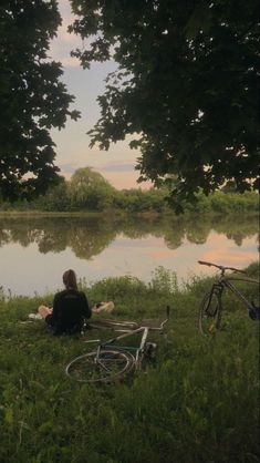 a woman sitting on the grass next to a lake with her bike leaning against it