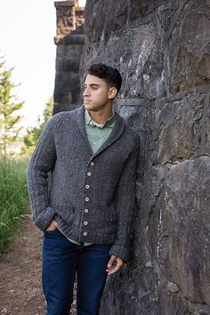 a man leaning against a stone wall wearing a sweater and jeans with his hands in his pockets