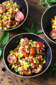 three bowls filled with corn and tomatoes on top of a wooden table next to green leaves