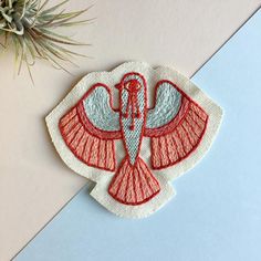 a red and white bird embroidered onto a piece of paper next to a succulent plant