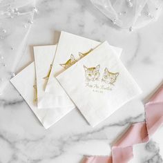 three white napkins with gold foil cats on them and pink ribbons around the edges