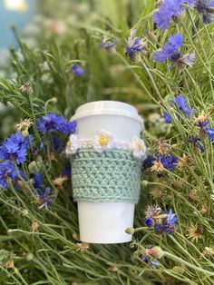 a crocheted coffee cup cozyie sits in the middle of some blue flowers