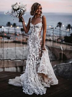 a woman in a white wedding dress holding flowers and smiling at the camera while standing on a wooden deck