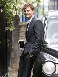 a man in a suit and tie standing next to a black car holding a drink