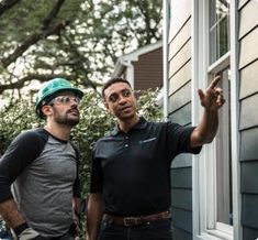 two men standing next to each other in front of a house and pointing at something