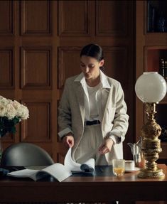 a woman standing at a desk in front of a book and papers with a lamp on it