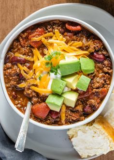 a white bowl filled with chili, cheese and avocado on top of a wooden table