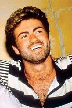 a man with a beard and striped shirt smiling in front of a cross on the wall