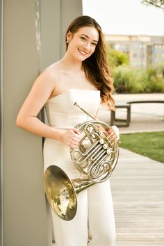 a woman in white is holding a french horn and smiling at the camera while standing on a porch