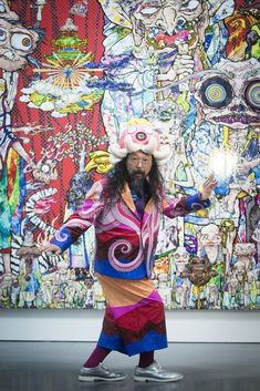 a man with long hair wearing a colorful outfit and hat standing in front of a large painting