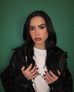 a woman with long black hair wearing a leather jacket and holding her hands on her chest