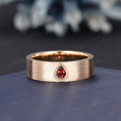 a gold wedding band with a pear shaped ruby stone in the center, on top of a blue surface