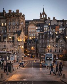 people are walking down the street in front of some old buildings at dusk with lights on