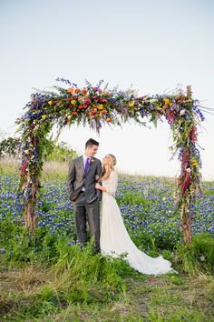 a bride and groom standing under an arch made out of flowers in the middle of a field