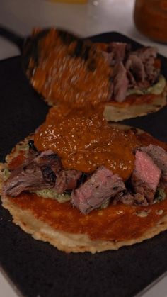 two pizzas with meat and sauce on them sitting on a black plate, ready to be eaten