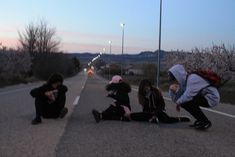 three people sitting on the side of a road with one person kneeling down and another standing up