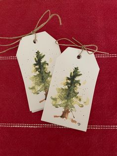 two tags with watercolor trees on them sitting on a red cloth covered tablecloth